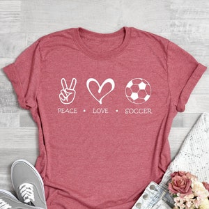 May include: A red t-shirt with a white peace sign, heart, and soccer ball graphic. The text "Peace Love Soccer" is printed below the graphic.