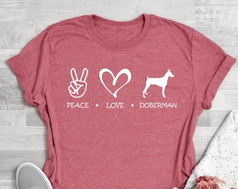 May include: Coral pink t-shirt with a white peace sign, heart, and Doberman silhouette. The text reads "Peace Love Doberman".