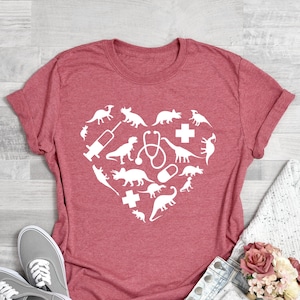 Puede incluir: Camiseta rosa coral con un dise&ntilde;o de coraz&oacute;n blanco con dinosaurios y s&iacute;mbolos m&eacute;dicos.