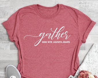 Könnte beinhalten: Korallenfarbenes T-Shirt mit dem Wort "gather" in Schreibschrift und dem Satz "here with grateful hearts" darunter. Das Shirt wird mit grauen Turnschuhen, Jeansshorts und einem weißen Pullover kombiniert. Ein kleiner Strauß rosa Blumen ist ebenfalls vorhanden.