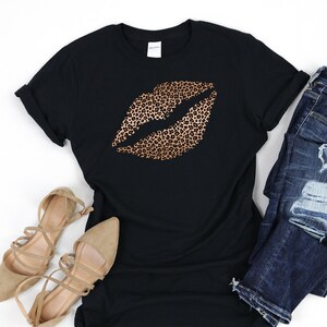 May include: Black t-shirt with a gold leopard print lip design. The shirt is paired with distressed blue jeans and brown strappy sandals.