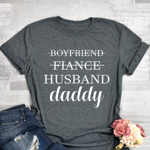 Puede incluir: Una camiseta gris con el texto "Boyfriend, Fiance, Husband, Daddy" impreso en blanco.