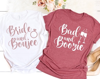 Op de afbeelding: Twee witte T-shirts met bedrukte tekst. Het linker shirt is wit met de tekst "Bride and Boujee" in roze schrift met een diamant-ring-afbeelding. Het rechter shirt is bordeauxrood met de tekst "Bad and Boozie" in wit schrift met een afbeelding van twee champagne glazen.