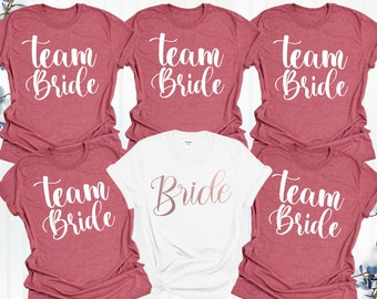 May include: Six t-shirts with the text "Team Bride" in white lettering on a maroon background. One white t-shirt has the text "Bride" in rose gold lettering.