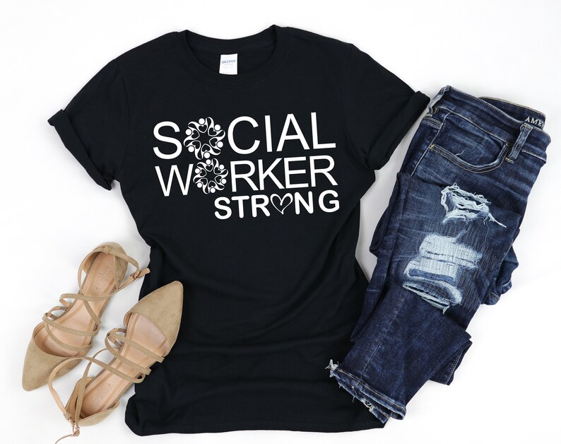 May include: Black t-shirt with white text that reads "SOCIAL WORKER STRONG" with a heart symbol in the word "STRONG". The shirt is paired with blue jeans and brown sandals.