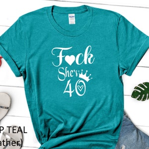 May include: Teal blue t-shirt with white text that says "F*ck She's 40" with a heart and crown graphic.