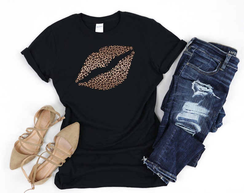 May include: Black t-shirt with a gold leopard print lip design. The shirt is paired with distressed blue jeans and brown strappy sandals.
