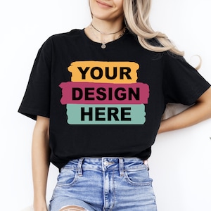 May include: Black t-shirt with the text "YOUR DESIGN HERE" in a bold, sans-serif font. The text is layered over three horizontal brush strokes in orange, magenta, and teal. The model is wearing blue jeans.