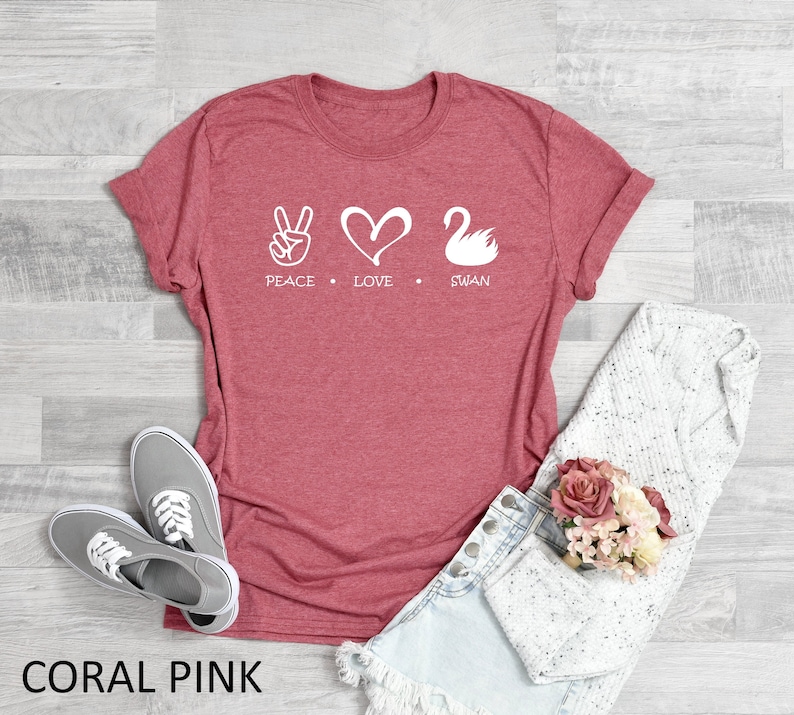 Puede incluir: Camiseta rosa coral con un gr&aacute;fico blanco de signo de paz, coraz&oacute;n y cisne. El texto "Peace Love Swan" est&aacute; impreso debajo del gr&aacute;fico.
