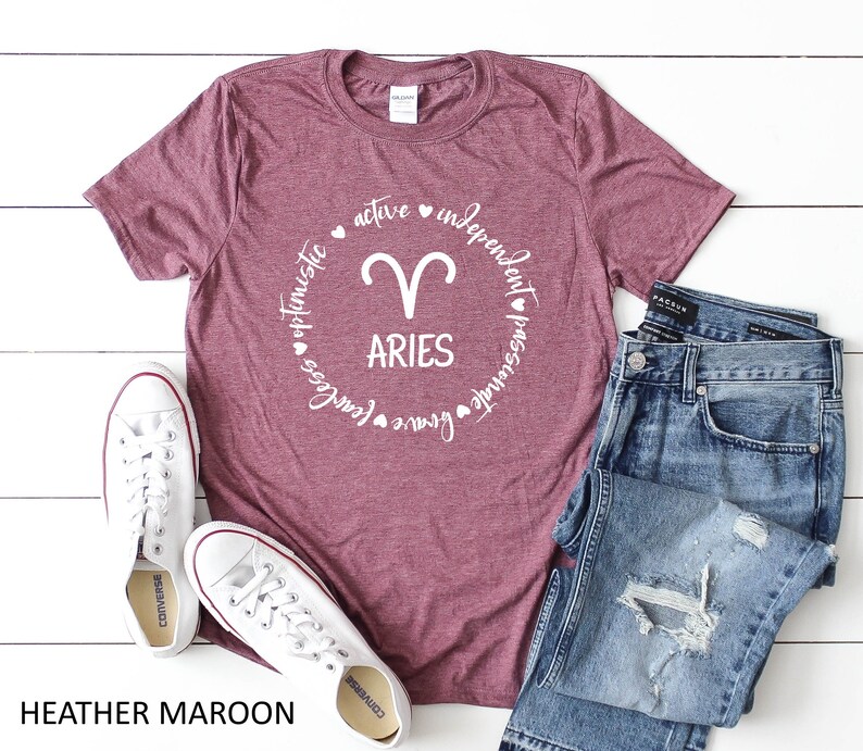 May include: Heather maroon t-shirt with a white Aries zodiac symbol and the word "ARIES" in a circle of descriptive words. The shirt is paired with blue jeans and white sneakers.