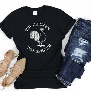 May include: Black t-shirt with a white graphic of a rooster and the text "The Chicken Whisperer". The shirt is paired with blue jeans and brown sandals.