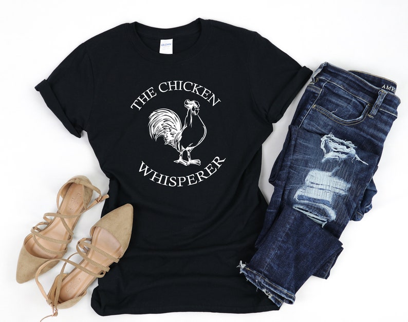 May include: Black t-shirt with a white graphic of a rooster and the text "The Chicken Whisperer". The shirt is paired with blue jeans and brown sandals.