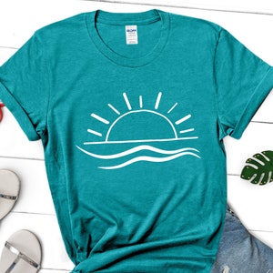May include: Teal blue t-shirt with a white graphic of a sun setting over waves.