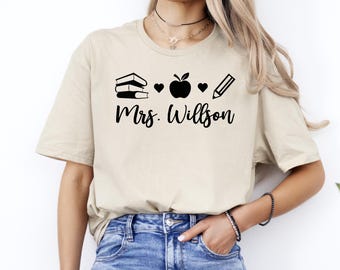 May include: A beige t-shirt with a black graphic design of a stack of books, two hearts, an apple, and a pencil. The text "Mrs. Wilson" is printed below the graphic.