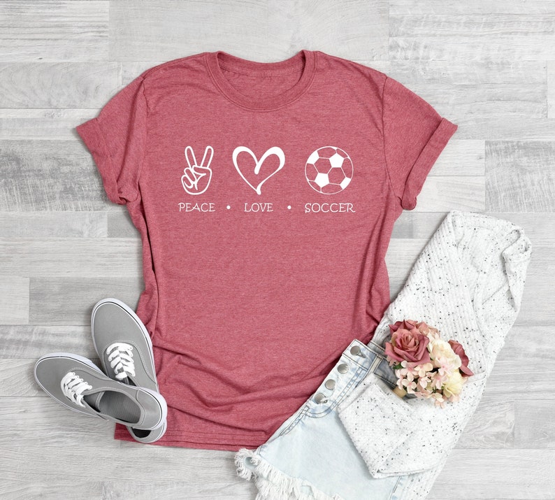May include: A red t-shirt with a white peace sign, heart, and soccer ball graphic. The text "Peace Love Soccer" is printed below the graphic.