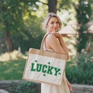 May include: A beige tote bag with a white front panel featuring the word "LUCKY" in green block letters, accompanied by green shamrock graphics. The bag has a natural jute-like texture and is carried by a person.
