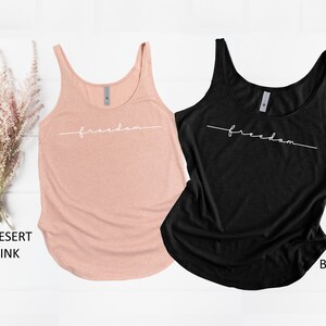 May include: Two tank tops with the word "freedom" written in white script on the front. The tank top on the left is a light pink color and the tank top on the right is black.