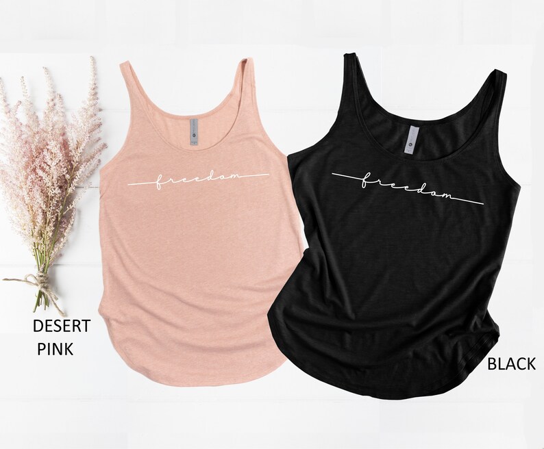 May include: Two tank tops with the word "freedom" written in white script on the front. The tank top on the left is a light pink color and the tank top on the right is black.