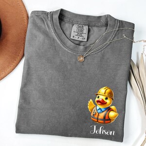 May include: A charcoal gray crew neck t-shirt featuring a graphic of a yellow rubber duck wearing a construction hard hat and an orange safety vest. The text "Johnson" is printed in white script below the duck. The shirt has a Comfort Colors label.