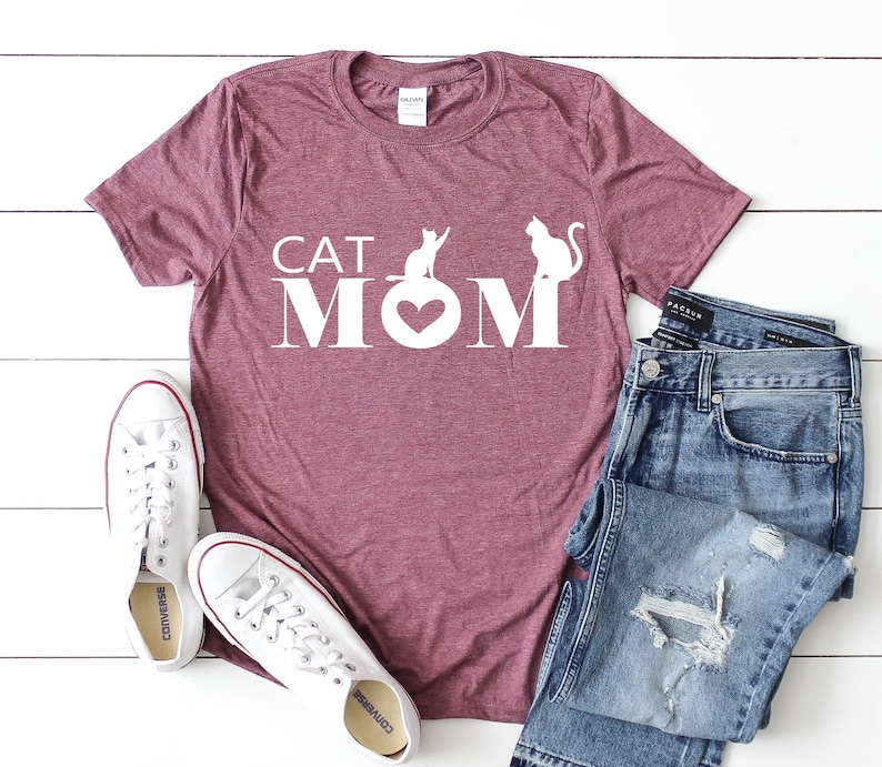 May include: A maroon t-shirt with the text "CAT MOM" in white. The "O" in "MOM" is a heart shape. Two white cat silhouettes are above the text. White sneakers and blue jeans are next to the shirt.