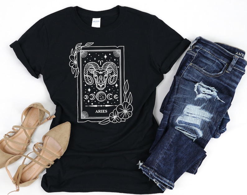 May include: Black t-shirt featuring a white graphic of the Aries zodiac sign. The design includes a ram's head, crescent moons, stars, and floral accents. The shirt is paired with blue denim jeans and beige sandals.