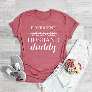Puede incluir: Una camiseta roja con el texto "Boyfriend - Fiance - Husband - Daddy" impreso en blanco.