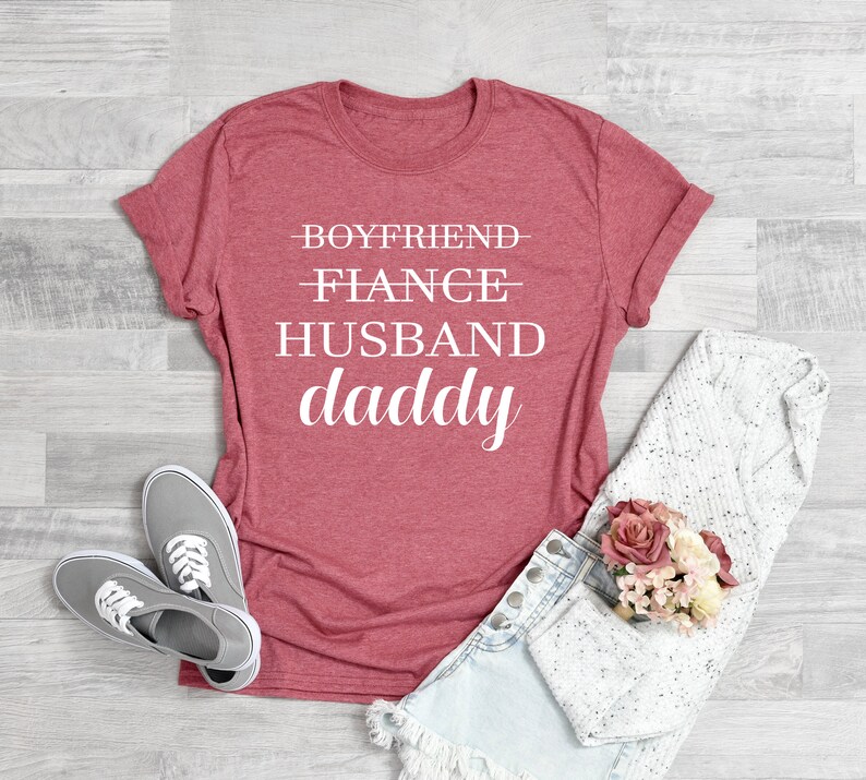 Puede incluir: Una camiseta roja con el texto "Boyfriend - Fiance - Husband - Daddy" impreso en blanco.
