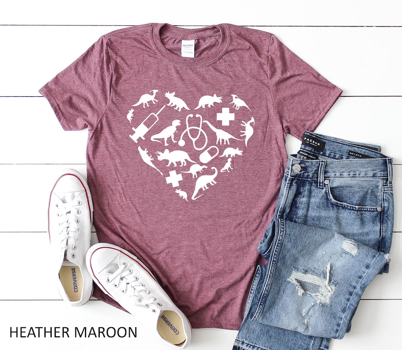 Puede incluir: Una camiseta de color marr&oacute;n rojizo con un gr&aacute;fico blanco de un coraz&oacute;n formado por dinosaurios y s&iacute;mbolos veterinarios. La camiseta est&aacute; sobre una superficie blanca con un par de zapatillas blancas y unos vaqueros azules.