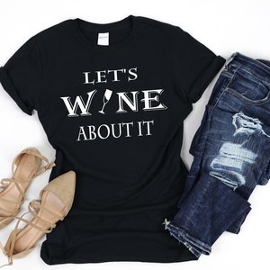 May include: Black t-shirt with white text that reads "Let's Wine About It" with a wine glass graphic. The shirt is paired with blue jeans and brown sandals.