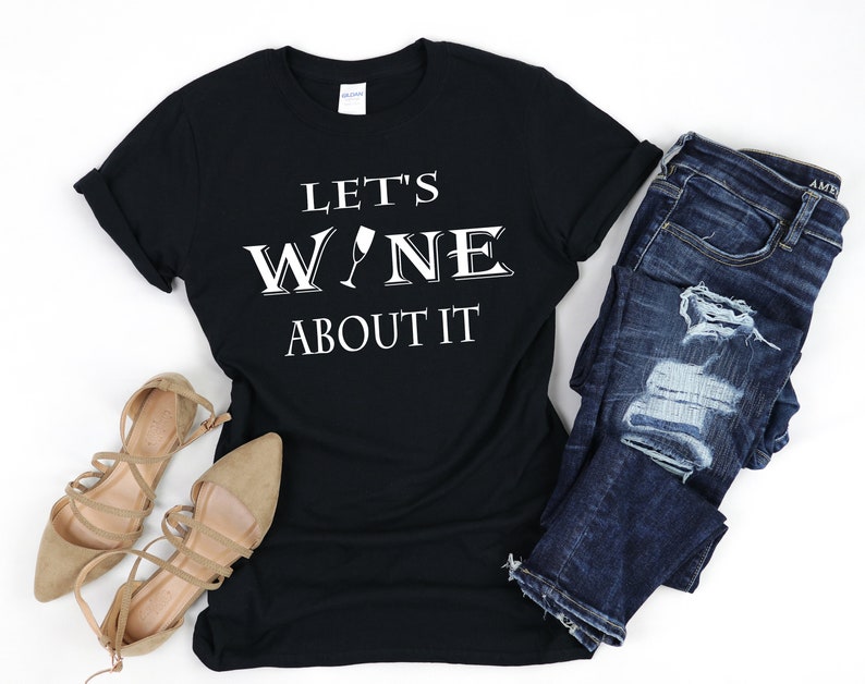 May include: Black t-shirt with white text that reads "Let's Wine About It" with a wine glass graphic. The shirt is paired with blue jeans and brown sandals.