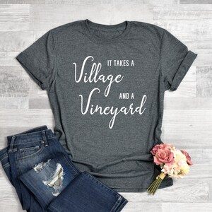 K&ouml;nnte beinhalten: Ein graues T-Shirt mit dem Text "Es braucht ein Dorf und ein Weingut" in wei&szlig;er Kursivschrift.