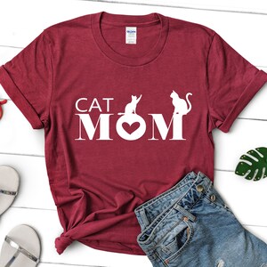 May include: A maroon t-shirt with the words "CAT MOM" in white block letters. The "O" in MOM is replaced with a heart shape. Two white cat silhouettes are positioned above the letters. The shirt is folded and styled with accessories.