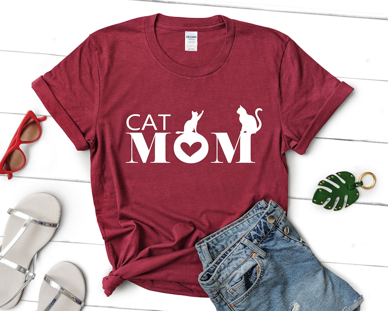 May include: A maroon t-shirt with the words "CAT MOM" in white block letters. The "O" in MOM is replaced with a heart shape. Two white cat silhouettes are positioned above the letters. The shirt is folded and styled with accessories.