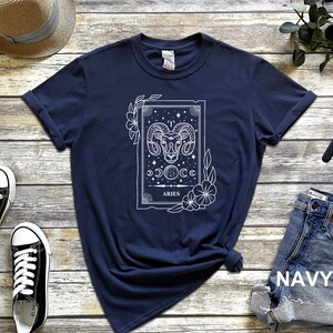 May include: Navy blue t-shirt featuring a white graphic of the Aries zodiac sign. The design includes a ram's head, celestial elements, and the word "ARIES". The shirt is styled with rolled-up sleeves and a knotted hem, showcasing a casual aesthetic.