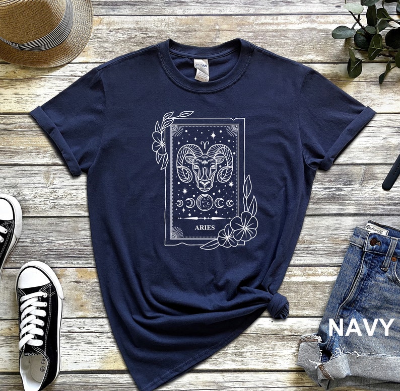 May include: Navy blue t-shirt featuring a white graphic of the Aries zodiac sign. The design includes a ram's head, celestial elements, and the word "ARIES". The shirt is styled with rolled-up sleeves and a knotted hem, showcasing a casual aesthetic.