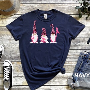 May include: Navy blue t-shirt with three gnomes wearing pink leopard print hats and holding pink ribbons. The gnomes are standing in a row.