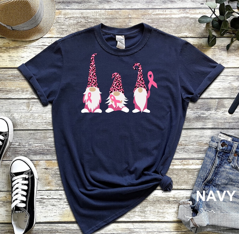 May include: Navy blue t-shirt with three gnomes wearing pink leopard print hats and holding pink ribbons. The gnomes are standing in a row.