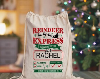 May include: A cream-colored drawstring gift bag with the text "REINDEER EXPRESS" in red and green. The bag is personalized "for RACHEL" and includes a checklist for "NAUGHTY" and "NICE". The bag is decorated with snowflakes and a reindeer sleigh design.