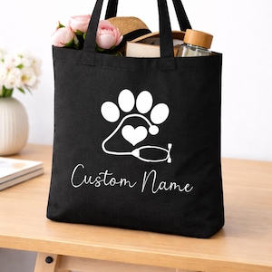 May include: A black tote bag featuring a white graphic of a paw print combined with a stethoscope and a heart. Below the graphic, the text Custom Name is printed in a white script font. The bag has two black shoulder straps and is filled with items.