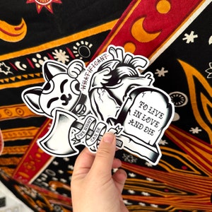 May include: Three black and white stickers featuring a heart, a tombstone, and a cat holding an axe. The tombstone reads "To live in love and die". The cat sticker says "What if I can't" and "The night can be".