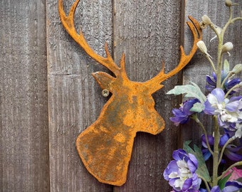 Puede incluir: Una silueta de cabeza de ciervo de metal oxidado montada en una pared de madera con flores rosas y moradas en primer plano.