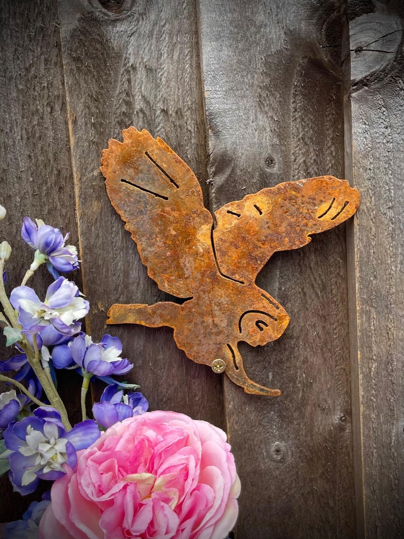 May include: A rusty metal owl silhouette with wings spread, mounted on a wooden surface. The owl is facing right and has a detailed design.