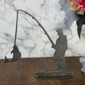 May include: Metal silhouette of a person fishing with a rod and a fish on the line. The silhouette is standing on a grassy base.