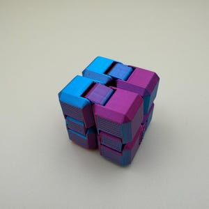 May include: A multi-jointed fidget cube constructed from interconnected plastic blocks. The object features a vibrant colour scheme with alternating bright blue and magenta purple segments, showcasing a textured, layered surface finish.