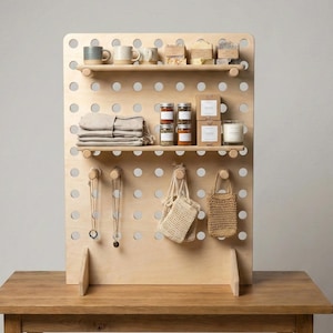 May include: A wooden display unit with a pegboard design, holding various items. There are mugs, jars, small boxes, folded linens, and candles on the shelves. Two necklaces and two small woven bags hang from the pegboard.