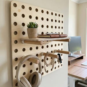 May include: A wooden pegboard organizer with multiple round holes, a shelf, and dowel rods for hanging items. It holds a small potted plant, a notebook, headphones, a charging cable, and keys. The pegboard is mounted on a beige wall.