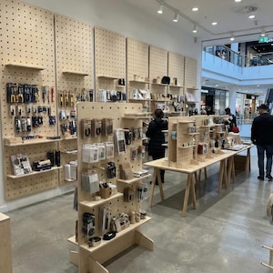 以下が含まれることがあります： 木製のディスプレイと有孔ボードの壁が特徴の小売店のインテリア。店内には、さまざまな工具、文房具、小さなアクセサリーが展示されています。照明は明るく、床はコンクリートです。顧客は商品を見ています。