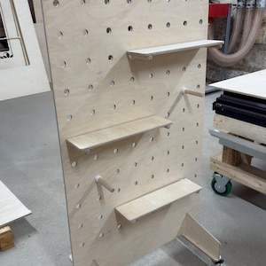 May include: A large, unfinished wooden pegboard with multiple shelves and dowel rods. The pegboard has evenly spaced holes for shelf and rod placement. The unit is supported by two legs with wheels, and is designed for display or storage.