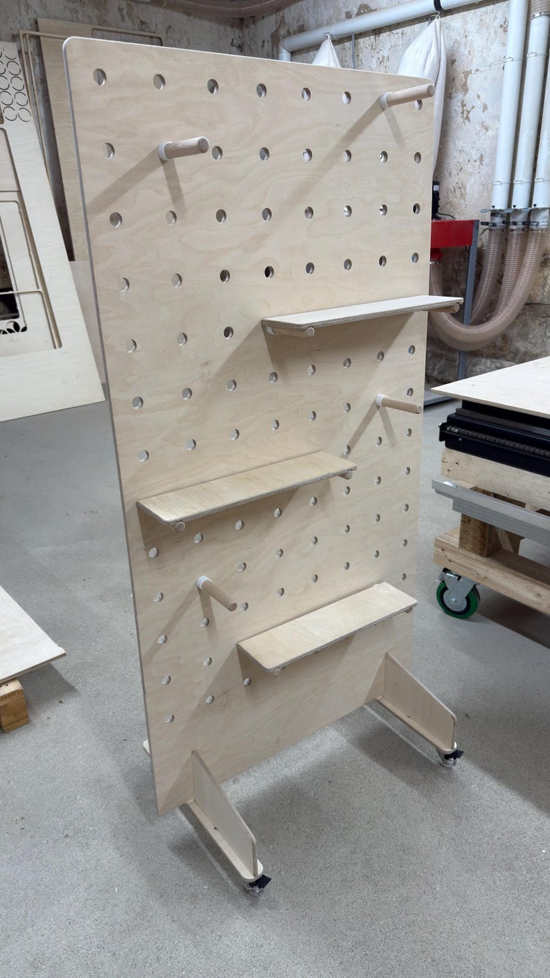 May include: A large, unfinished wooden pegboard with multiple shelves and dowel rods. The pegboard has evenly spaced holes for shelf and rod placement. The unit is supported by two legs with wheels, and is designed for display or storage.