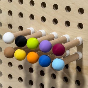 May include: A wooden pegboard featuring ten multi-coloured ball-shaped pegs. The pegs are white, black, green, purple, red, brown, yellow, orange, blue, and light blue. The pegs are attached to wooden dowels with white plastic supports.